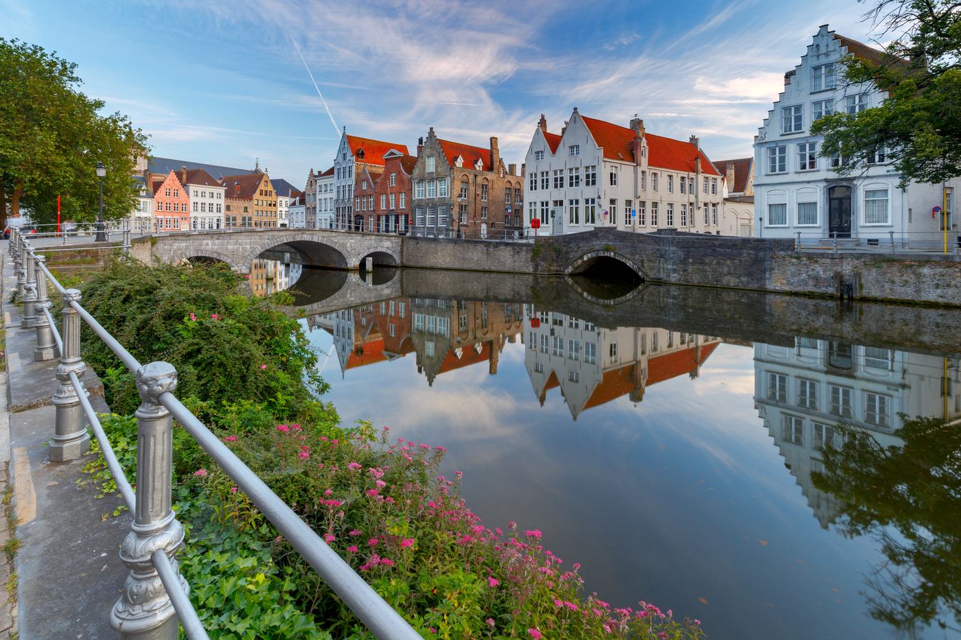 A calm Bruges canal with a stone bridge, pink flowers and tall gabled houses