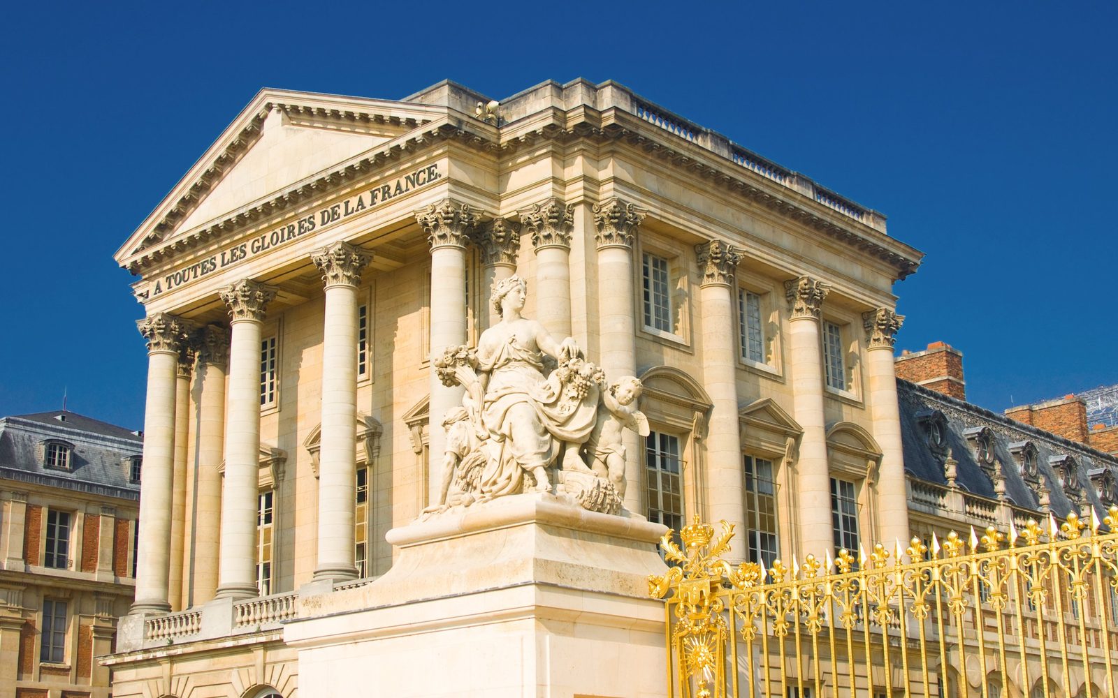 The Palace of Versailles facade with a stone statue and golden gate under a deep blue sky
