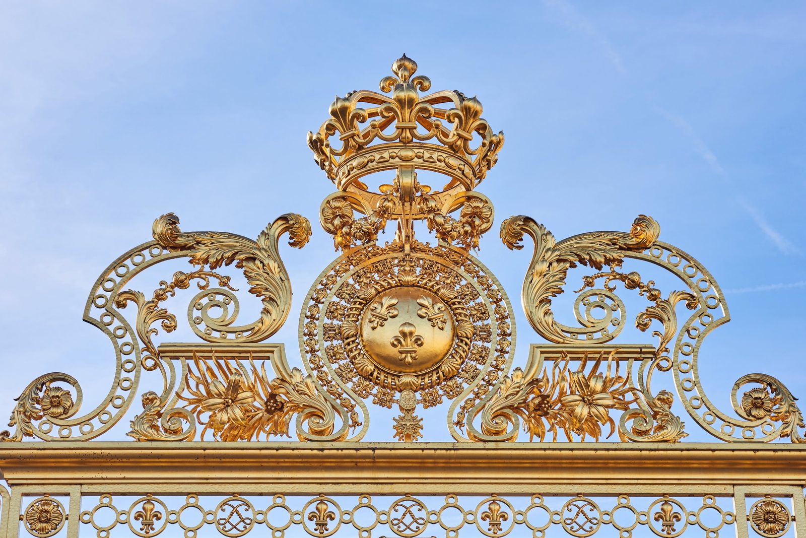 Gold crown and fleur-de-lis ornament on the royal gate of Versailles against a blue sky